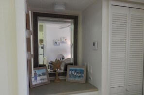 A room with a vanity area, a mirror, and a small shelf with framed pictures. There is a white door with louvered shutters to the right.