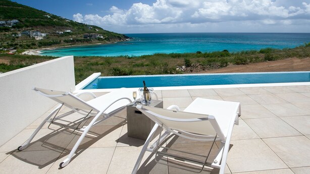 A terrace with a pool overlooking a coastal view, featuring a white lounge chair and a bottle of champagne. - Villa Amaya - Modern, Pristine Villa, Stunning Ocean Views, Private Pool, Beach (Indigo Bay)