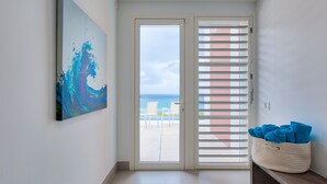 A room with a view of the sea through a sliding glass door, a painting on the wall, and a basket with towels on a bench. - Villa Amaya - Modern, Pristine Villa, Stunning Ocean Views, Private Pool, Beach (Indigo Bay)