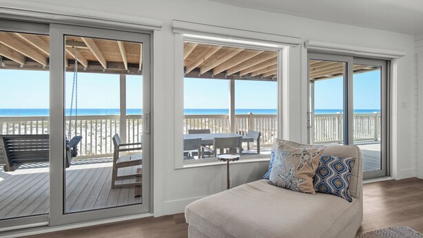 A room with a large sliding glass door leading to a wooden deck overlooking a beach.