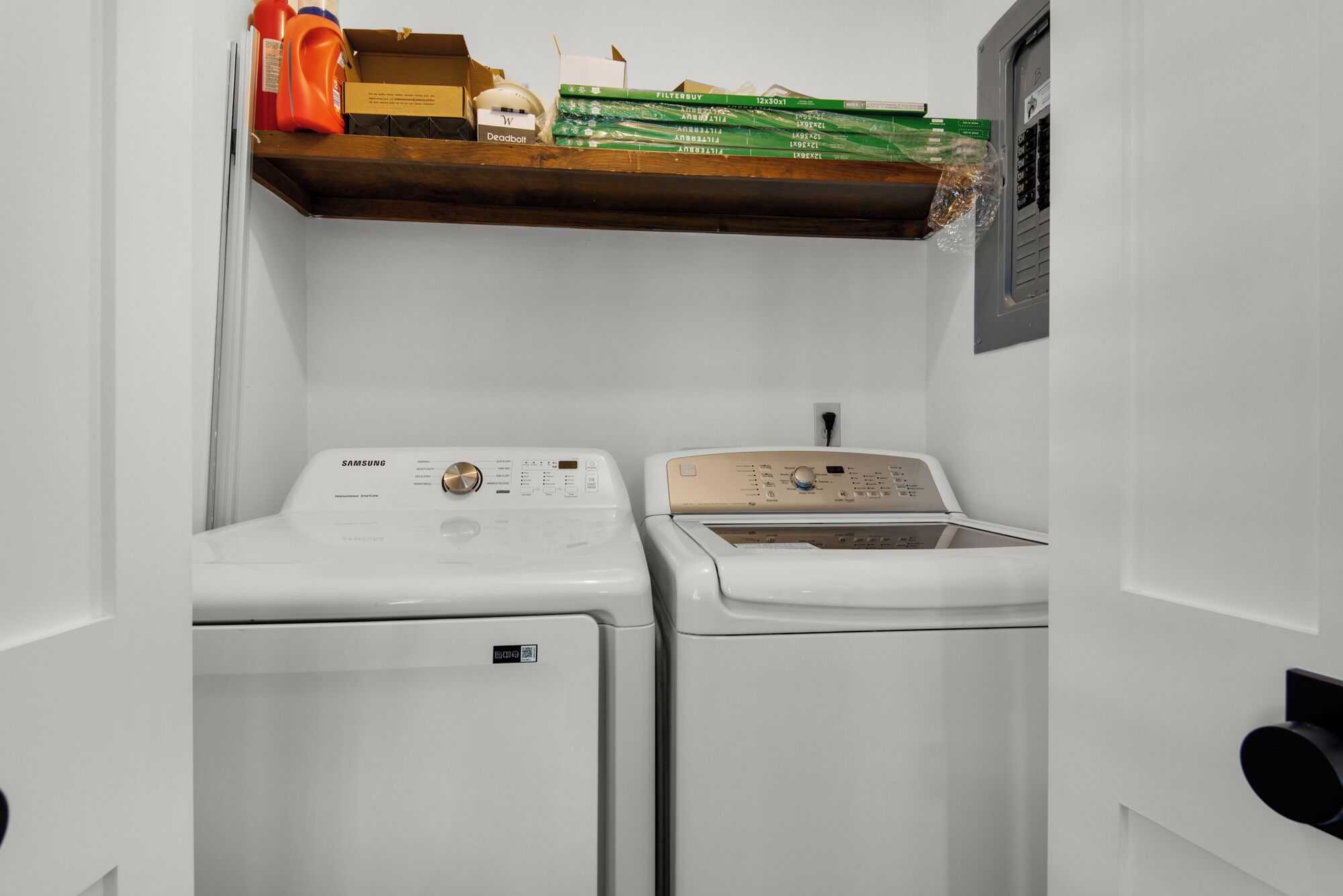 A laundry room with two white washing machines and a dryer, a wooden shelf above with various items, and a window with blinds.