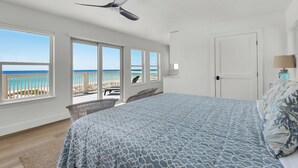 A bedroom with a large bed, a ceiling fan, and a view of the beach through large windows.