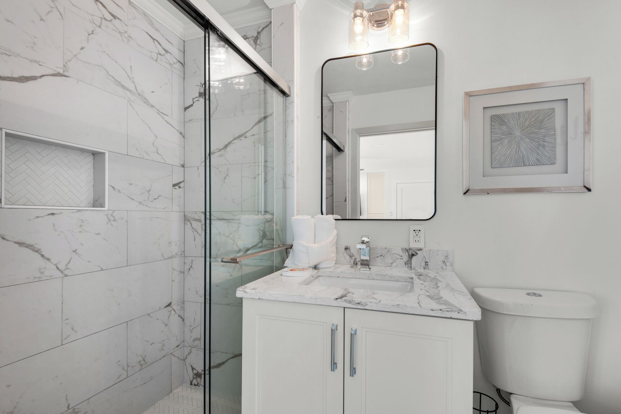 Un baño moderno con cabina de ducha de vidrio, pared con azulejos de mármol, lavamanos blanco con encimera de mármol y una obra de arte enmarcada sobre el espejo.