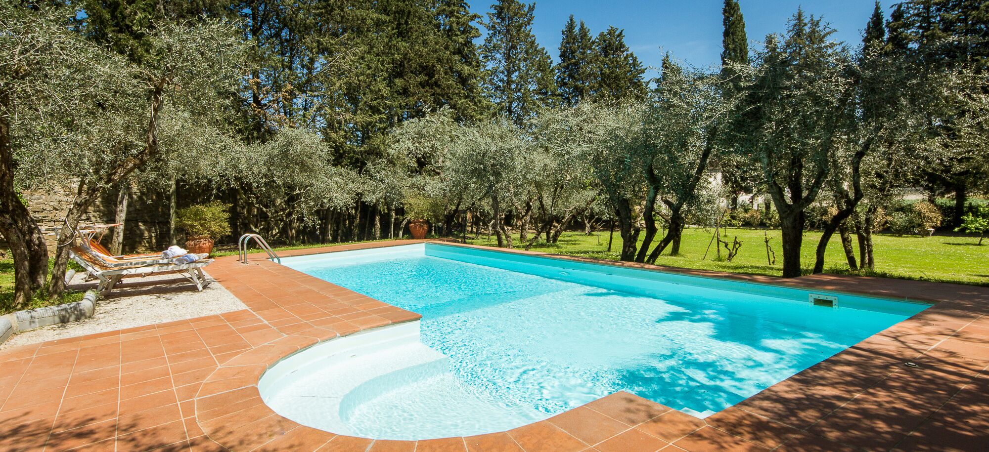 Une piscine rectangulaire, bordée d’une terrasse carrelée, entourée d’arbres et d’une pelouse.