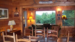 Interior d'una cabana amb parets de fusta, un sofà, seients, una taula de menjador i una finestra amb vista a arbres.