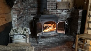 Una habitación rústica con una chimenea de ladrillo, un banco de madera y un cubo de metal lleno de periódicos.