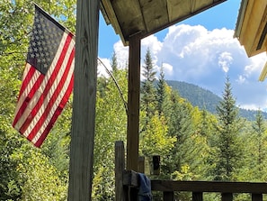 Una terraza de madera con una bandera estadounidense, rodeada de exuberante follaje y una montaña al fondo.