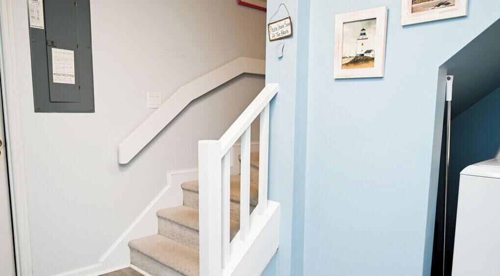 A staircase with a white handrail and beige carpeted steps, leading to a landing with a light blue wall and framed pictures.
