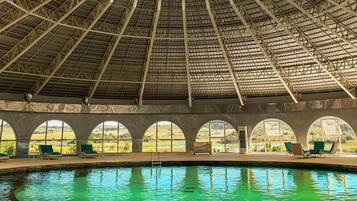 An indoor swimming pool with a glass ceiling, multiple arched windows, and lounge chairs along the poolside.