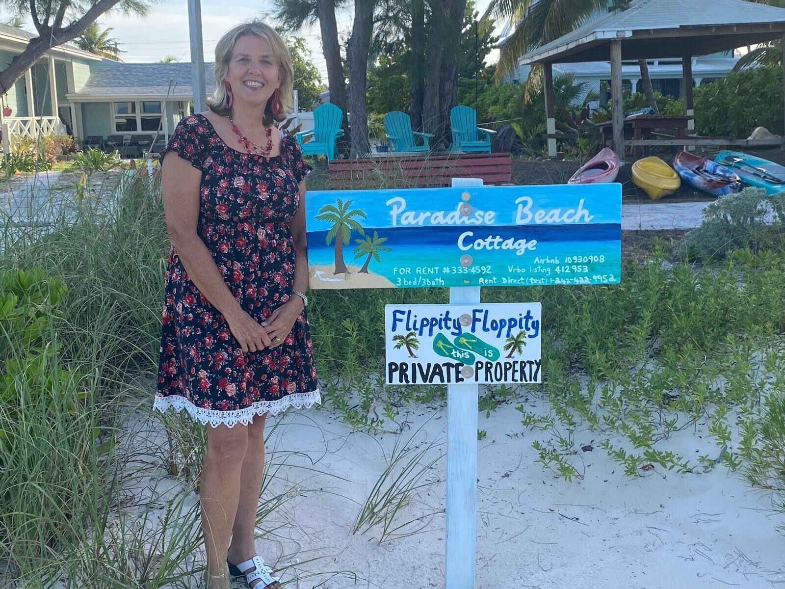A woman standing in front of a sign for Paradise Beach Cottage, which advertises a 3-bed/3-bath accommodation, with contact details for rent and direct text.