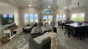 A spacious living room with a large TV, comfortable seating, a dining area, and large windows offering a view of the outdoors. - Paradise Beach Cottage- Newly Renovated 3 Bed/3 Bath Cottage On The Beach (Eleuthera)