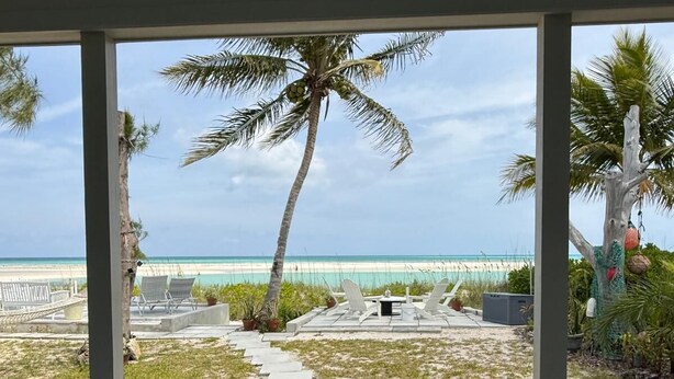 A covered outdoor area with a pathway leading to a beach, featuring palm trees, lounge chairs, and potted plants. - Paradise Beach Cottage- Newly Renovated 3 Bed/3 Bath Cottage On The Beach (Eleuthera)