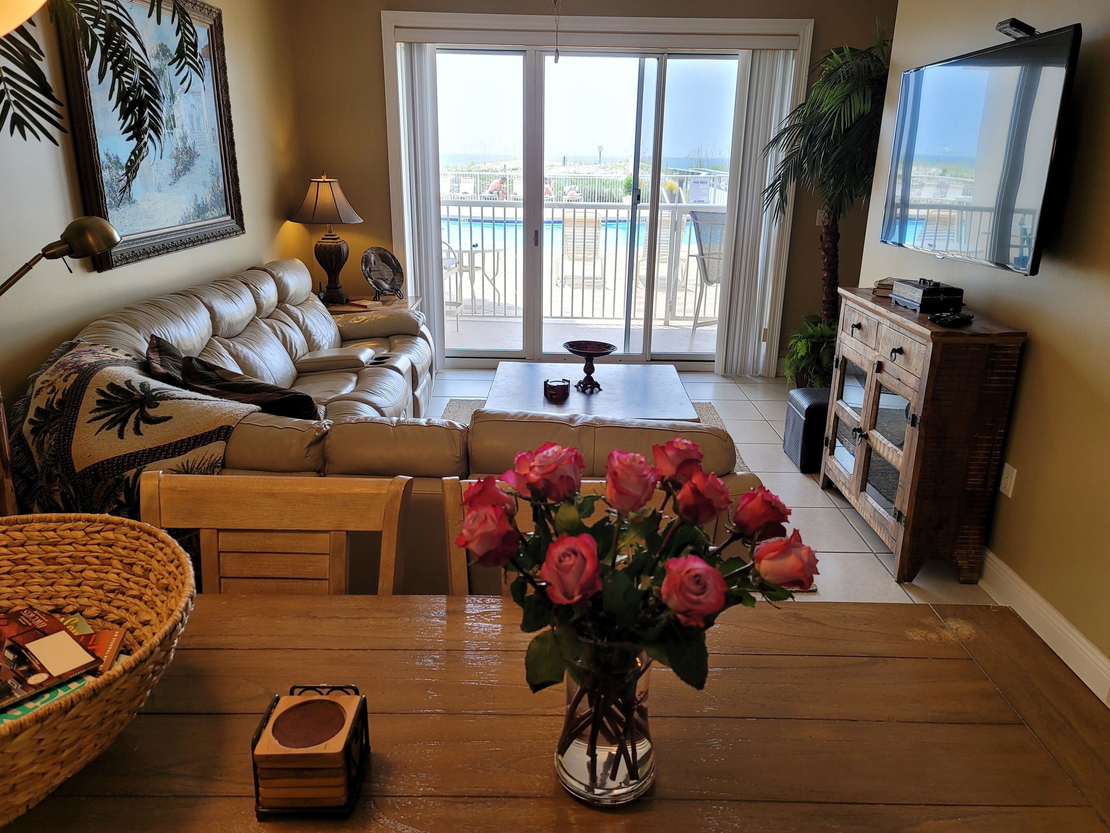A living room with a sofa, a coffee table, a TV, and a view of a pool and beach through a sliding door.