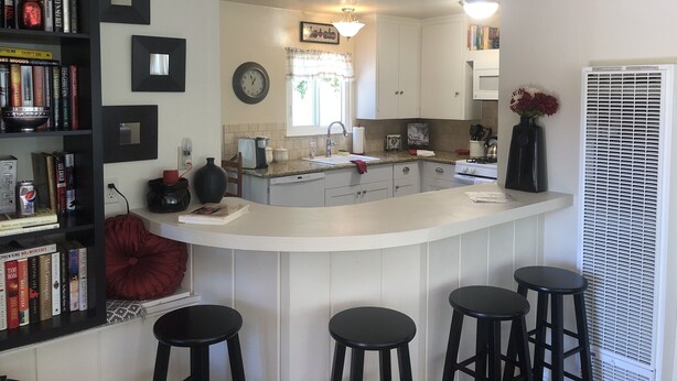 Una cocina con una isla central, taburetes de bar, una estantería y una ventana con persianas.