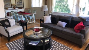 Un salón con un sofá gris, una mesa de centro redonda y una zona de comedor con mantel rojo.