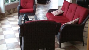 Un salon avec un sol à carreaux, deux canapés rouges, un fauteuil en osier noir, un ventilateur de plafond et un téléviseur sur le mur de droite.