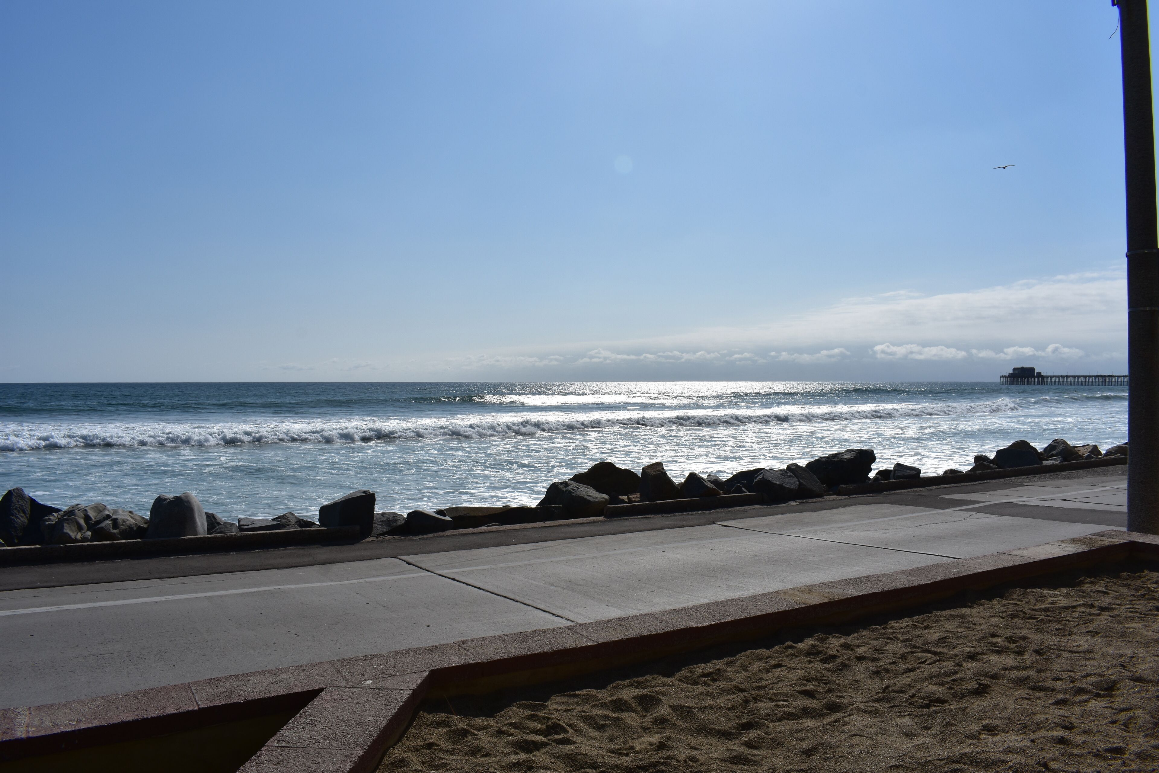 A coastal view with a concrete walkway, sandy beach, and a rocky shoreline.