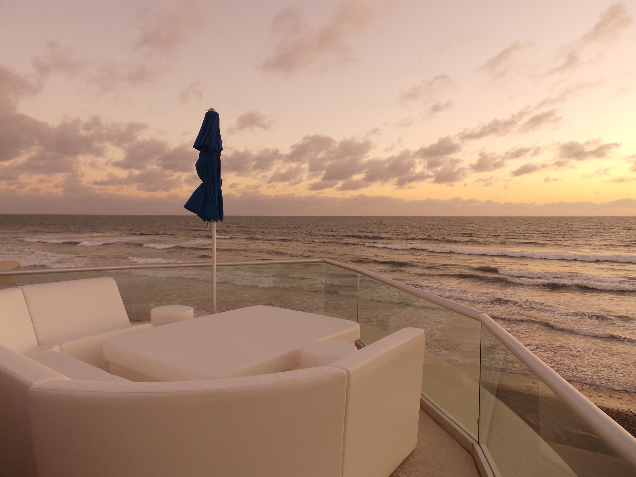 A balcony with white cushioned seating and a blue umbrella, overlooking the ocean at sunset.