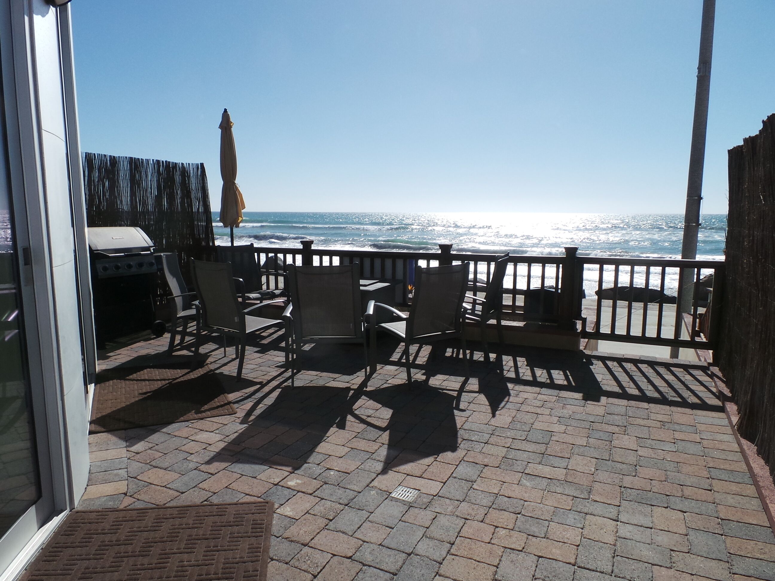 An outdoor patio with brick flooring, several chairs, a grill, and a view of the ocean.