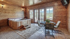 Interior de una cabaña de madera con jacuzzi, zona de estar y grandes ventanales que ofrecen vistas a la montaña.