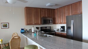 A kitchen with wooden cabinets, a stainless steel refrigerator, a microwave, and a sink. There are wicker chairs and a glass table. - Oceanfront 2 BR/2BA in Beautiful Sea Village. (Kailua-Kona)