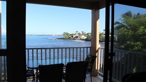 A balcony with a glass table and chairs overlooking a coastal view with palm trees and buildings. - Oceanfront 2 BR/2BA in Beautiful Sea Village. (Kailua-Kona)