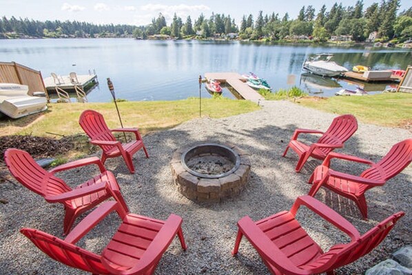 A lakeside deck with red Adirondack chairs, a fire pit, and boats docked in the background.