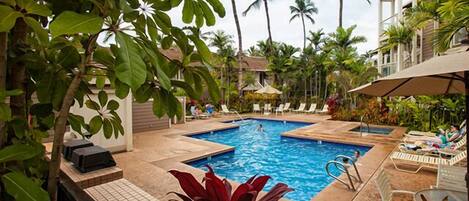 A pool area with lounge chairs and umbrellas, surrounded by palm trees and a building.