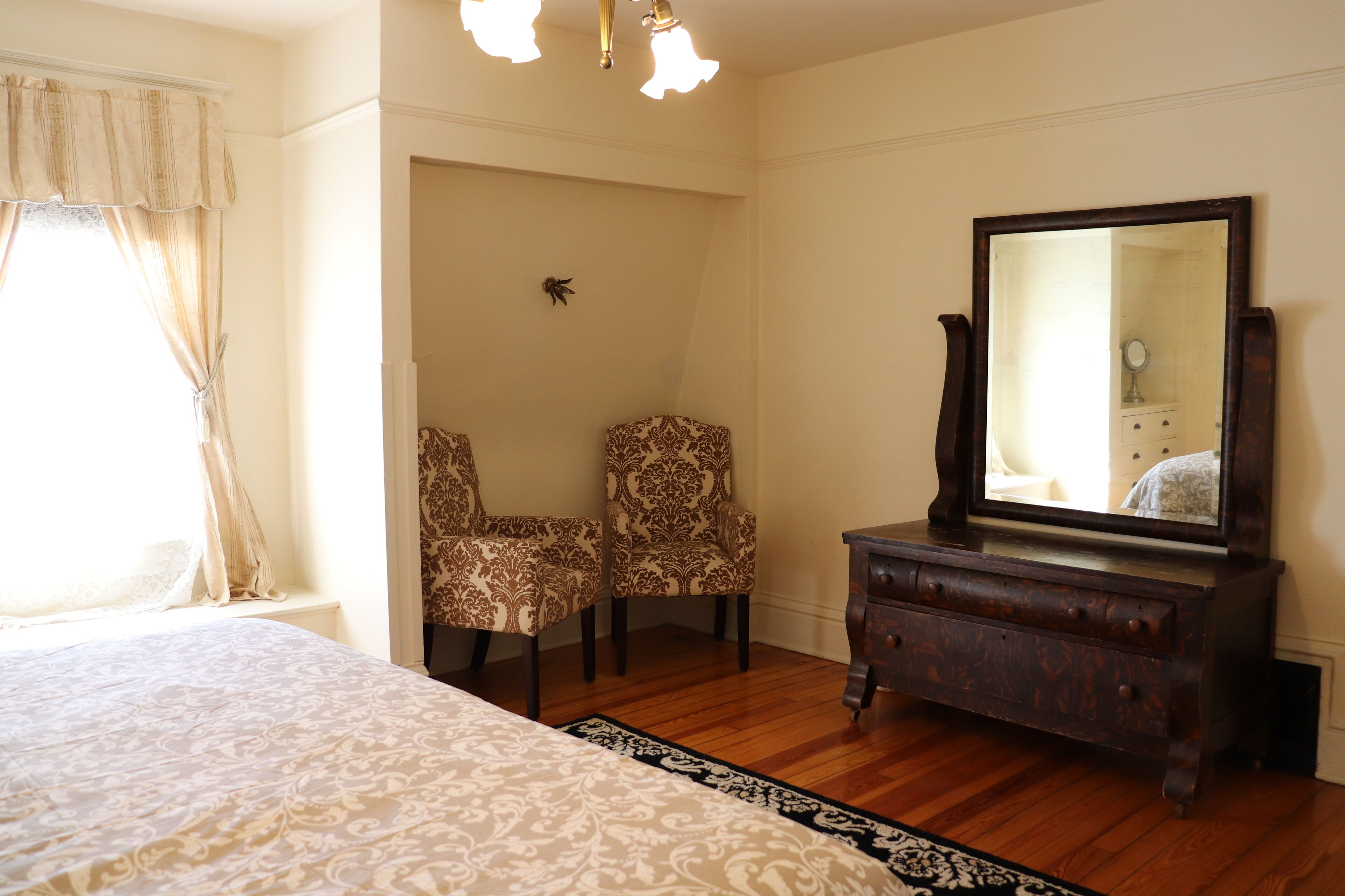 A bedroom with a bed, two patterned chairs, a dresser, and a large mirror.