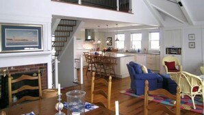 Ampla área de estar tipo loft, com teto alto, lareira, espaço para refeições e cozinha com grandes janelas.