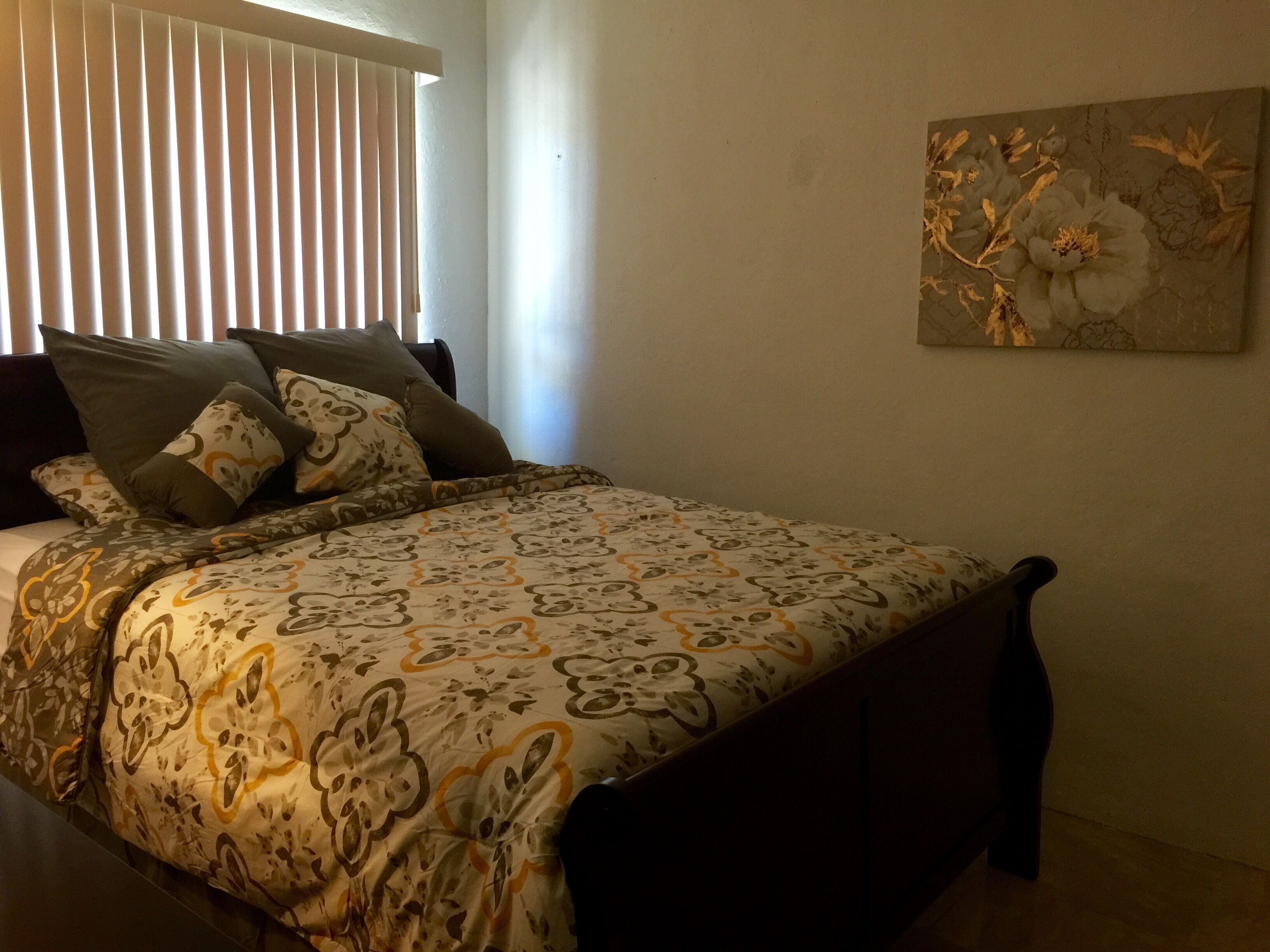 A bedroom with a bed, pillows, a window with blinds, and a floral painting on the wall.