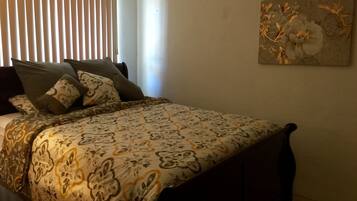 A bedroom with a bed, pillows, a window with blinds, and a floral painting on the wall.