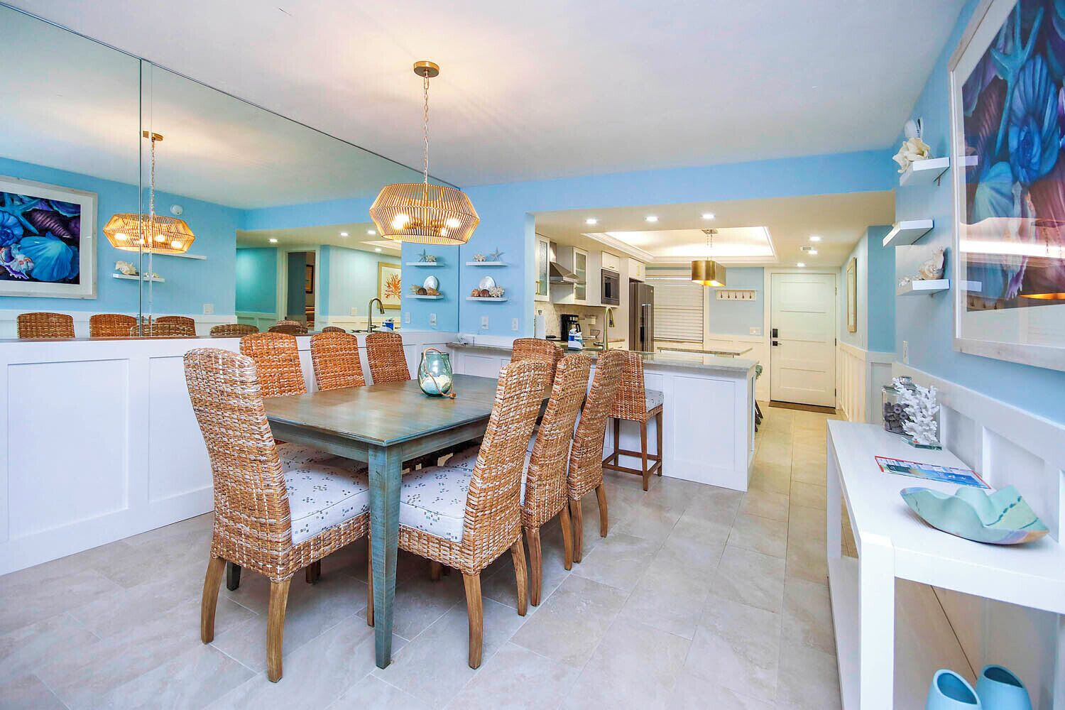 A dining area with a large table and wicker chairs, a kitchen in the background, and a blue wall with artwork.