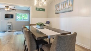 A dining area with a wooden table, grey upholstered chairs, a chandelier, a painting of sailboats, and a 'BEACH' sign on the wall. - Frances Cottage - Walking distance to downtown Saugatuck! Large backyard! (Saugatuck)