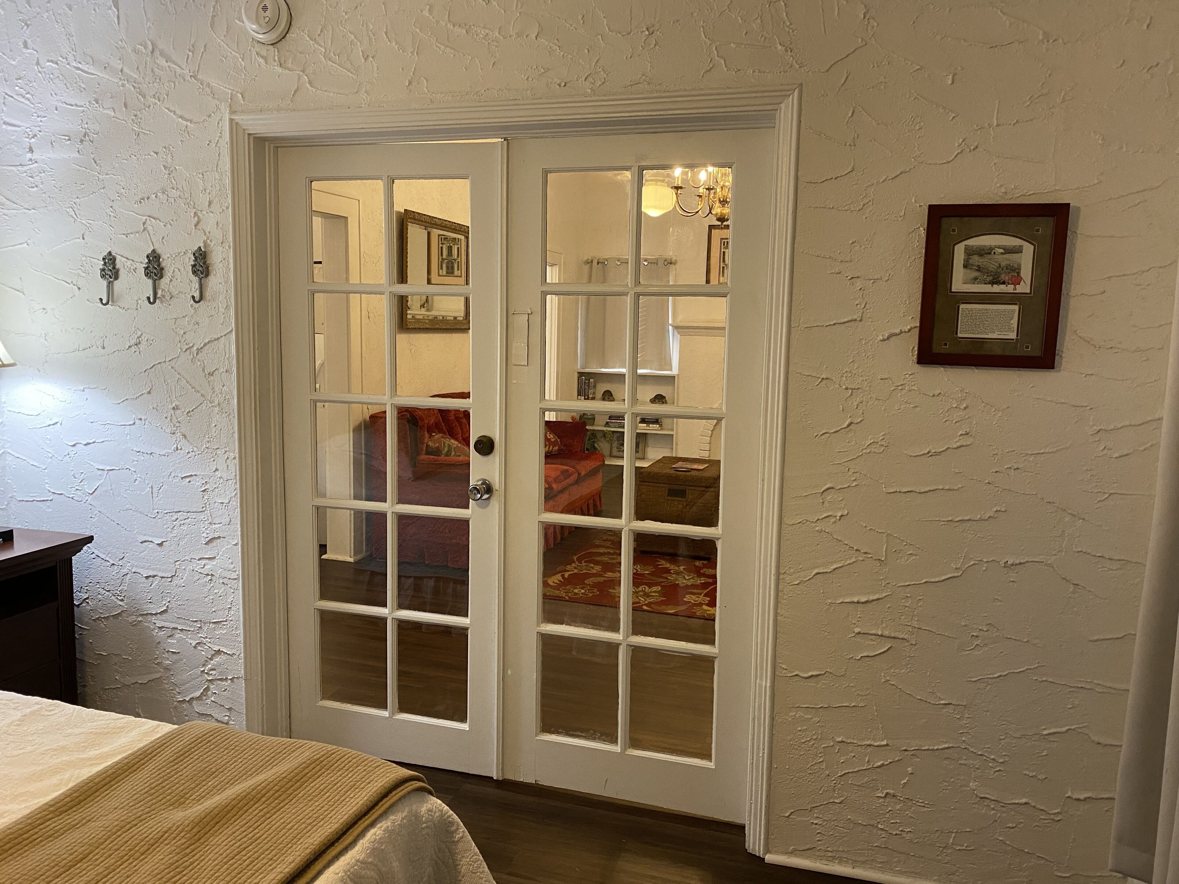 A room with a double door leading to another room, a bed with a beige cover, and a wall with a framed picture.