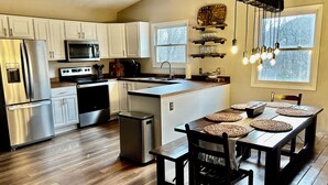A modern kitchen with white cabinets, stainless steel appliances, a wooden dining table with woven placemats, and a hanging light fixture. - Relaxing retreat on Otter Falls with views of Grandfather Mountain. (Seven Devils)