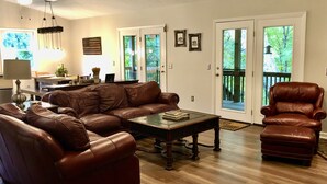 A living room with a brown leather sofa, a glass coffee table, and a brown leather armchair. There is a dining area with a table and chairs, and a kitchen in the background. - Relaxing retreat on Otter Falls with views of Grandfather Mountain. (Seven Devils)