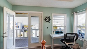 Una habitación con escritorio, una silla, ventana con persianas, puerta que da a un balcón y una decoración de timón de barco en la pared.