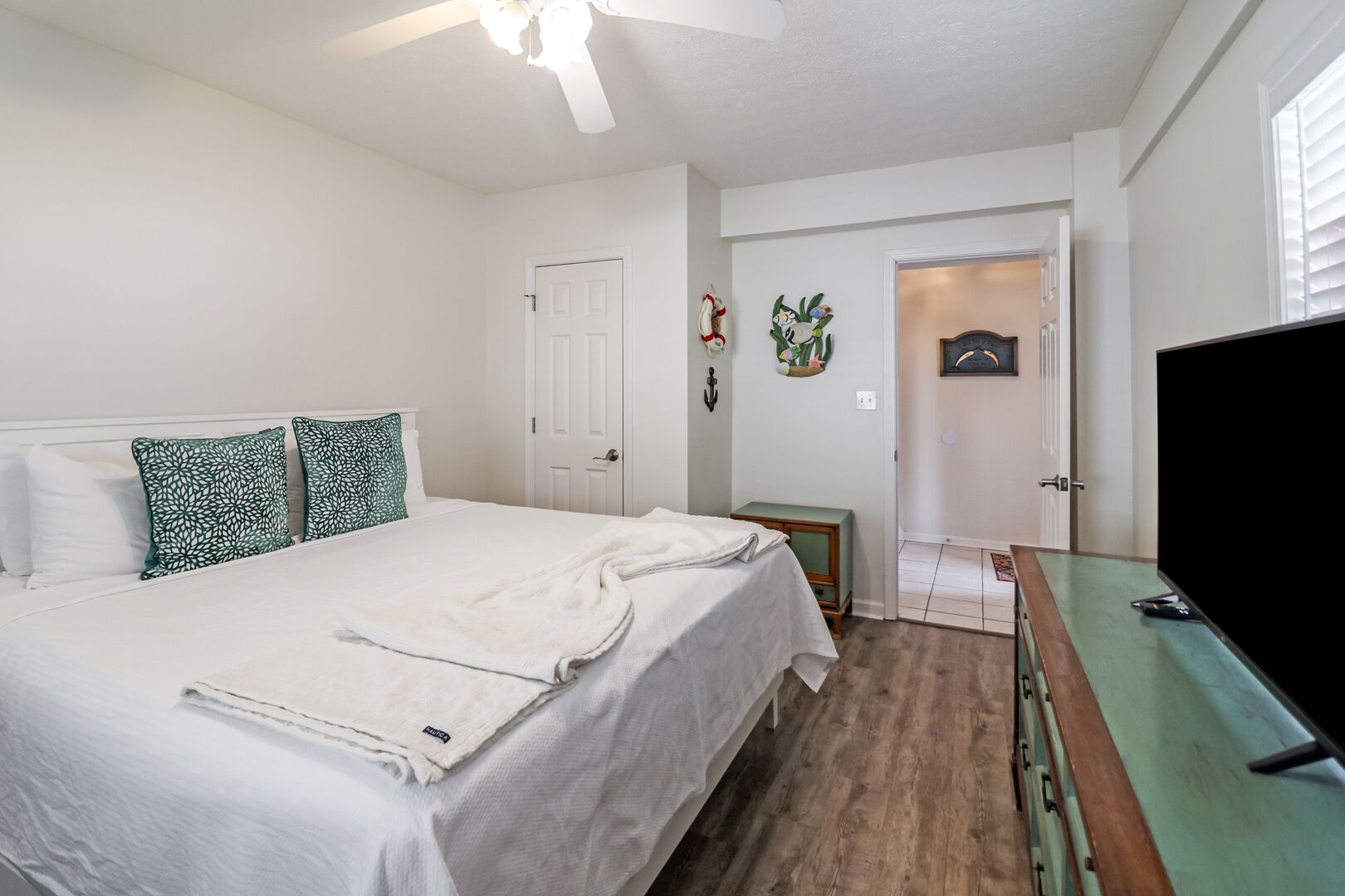 A bedroom with a bed, a TV, a ceiling fan, and a door leading to another room.