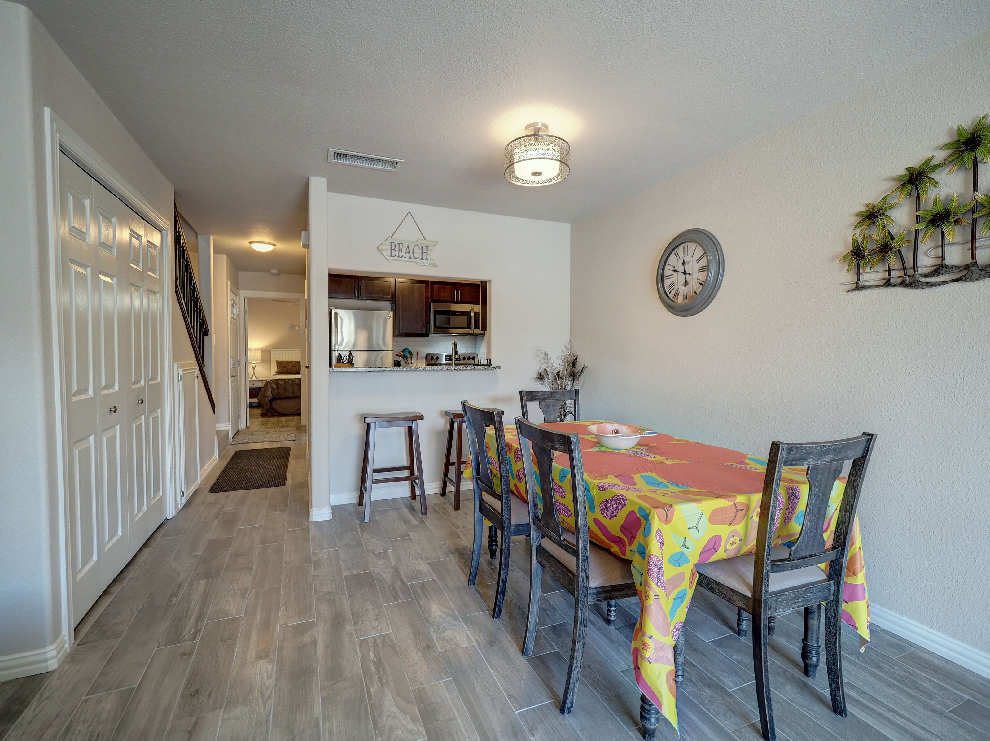 Uma área de refeições com uma mesa comprida, cadeiras, uma cozinha ao fundo e uma escada que leva a outro andar.
