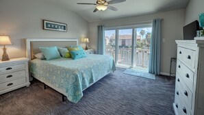 Quarto com cama, duas mesas de cabeceira, ventilador de teto, janela grande com vista para uma área residencial e cômoda branca.