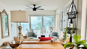 A spacious living room with a wooden table, two grey armchairs, a red cushion, a ceiling fan, and large windows offering a view of the outdoors.