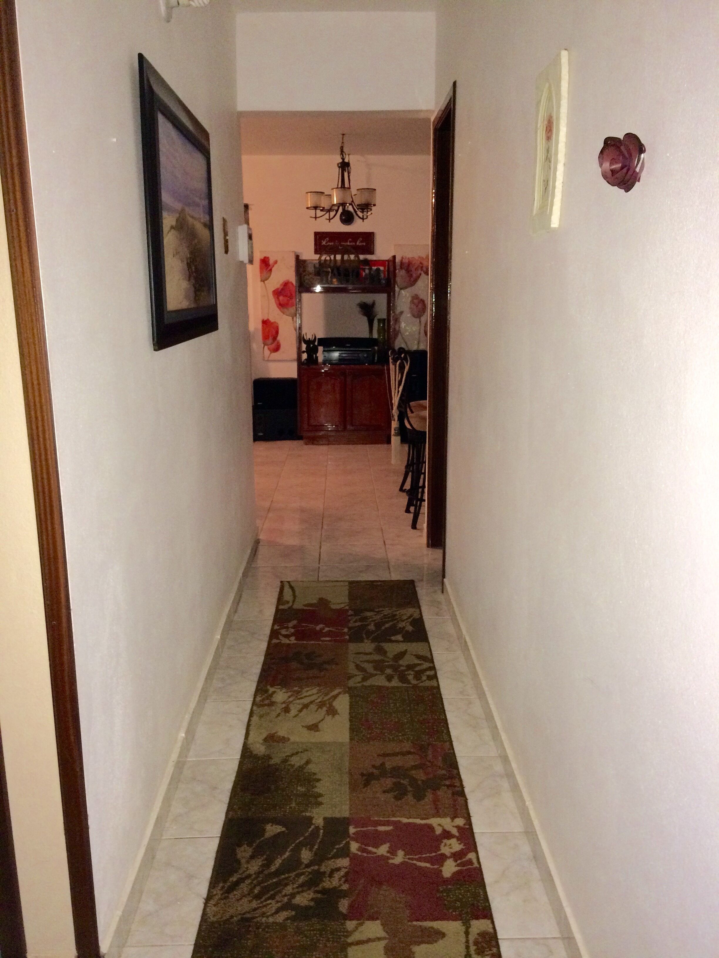 A hallway with a patterned rug, a chandelier, and a dining area in the background.