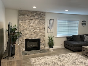 A living room with a stone fireplace, a flat-screen TV, a gray sofa, and a window with blinds. - Camp Pendleton Retreat with 7 person SPA 5bd 2 bath sleeps 12 (Oceanside)