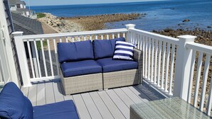 Une terrasse avec un canapé bleu et des coussins, une table à plateau en verre et une balustrade blanche donnant sur une plage.