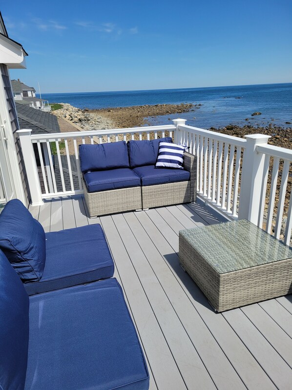 Una terraza con un sofá azul y cojines, una mesa con cubierta de vidrio y una barandilla blanca con vista a la playa.