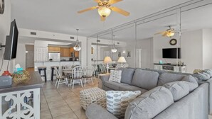 Un salon moderne avec un canapé gris, un coin repas avec des chaises blanches, une cuisine en arrière-plan et un ventilateur de plafond.