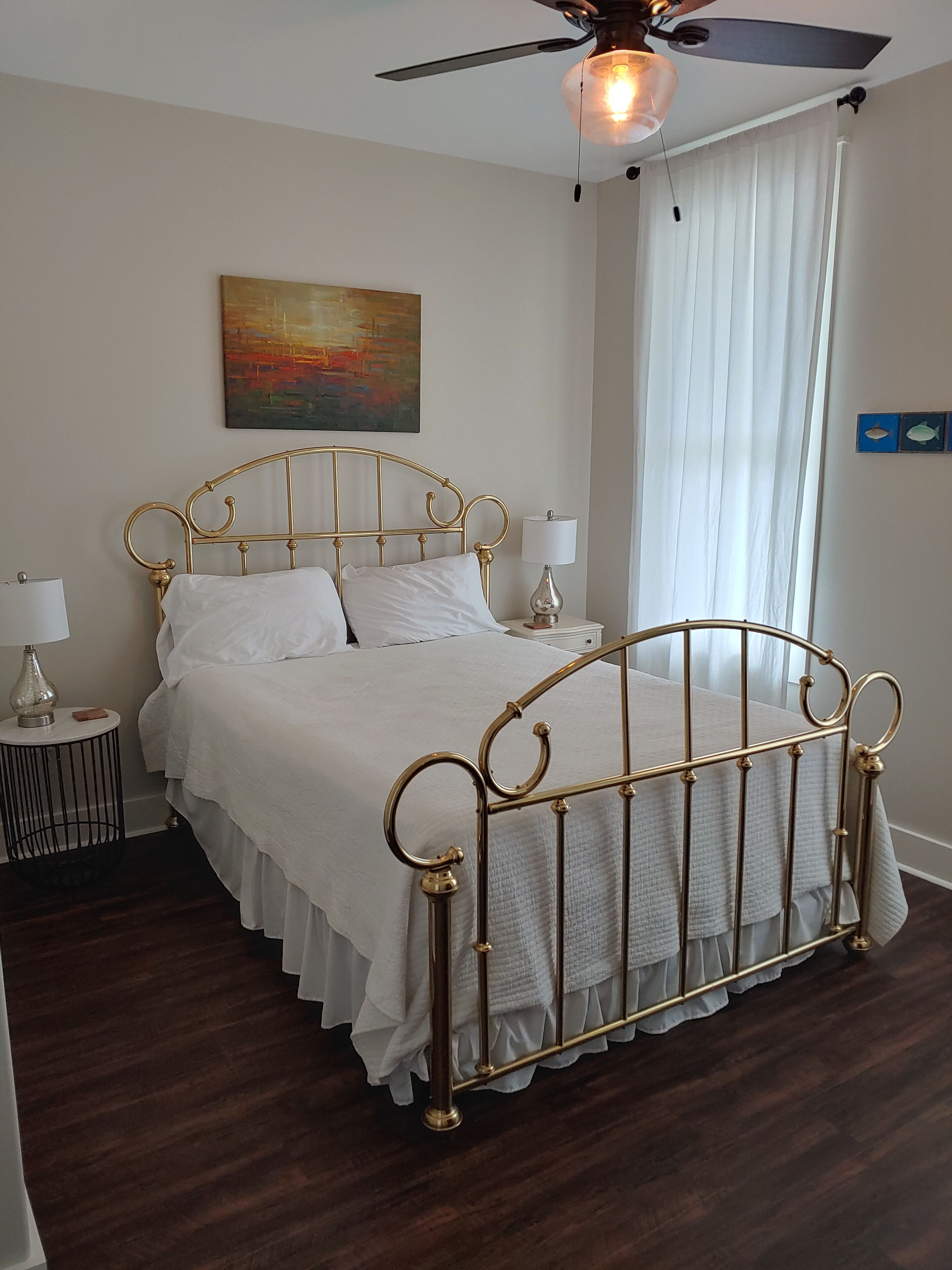 Une chambre à coucher avec un lit à ossature métallique, du linge de lit blanc, un ventilateur de plafond et un tableau accroché au mur.