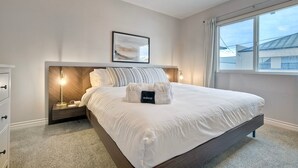 Uma cama bem arrumada com edredom branco e travesseiros, uma mesinha de cabeceira com abajur e um quadro na parede.