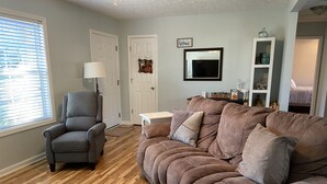 A living room with a grey armchair, a brown sofa with cushions, a white side table, and a wall-mounted TV. - Quiet, Cozy, and Family Friendly in Hendo (Hendersonville)